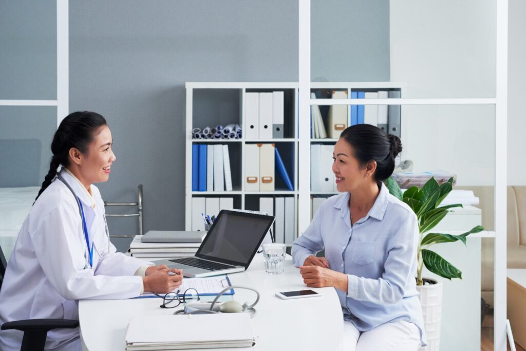 Two healthcare professionals in white coats sit at a desk with a laptop and documents, smiling as they talk in a bright clinic.